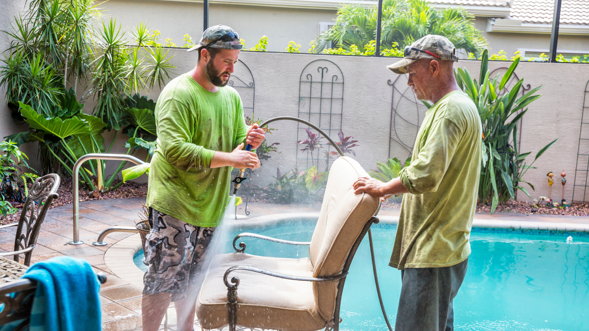 Two workers cleaning a patio chair by a swimming pool, surrounded by tropical plants, emphasizing outdoor maintenance and craftsmanship.