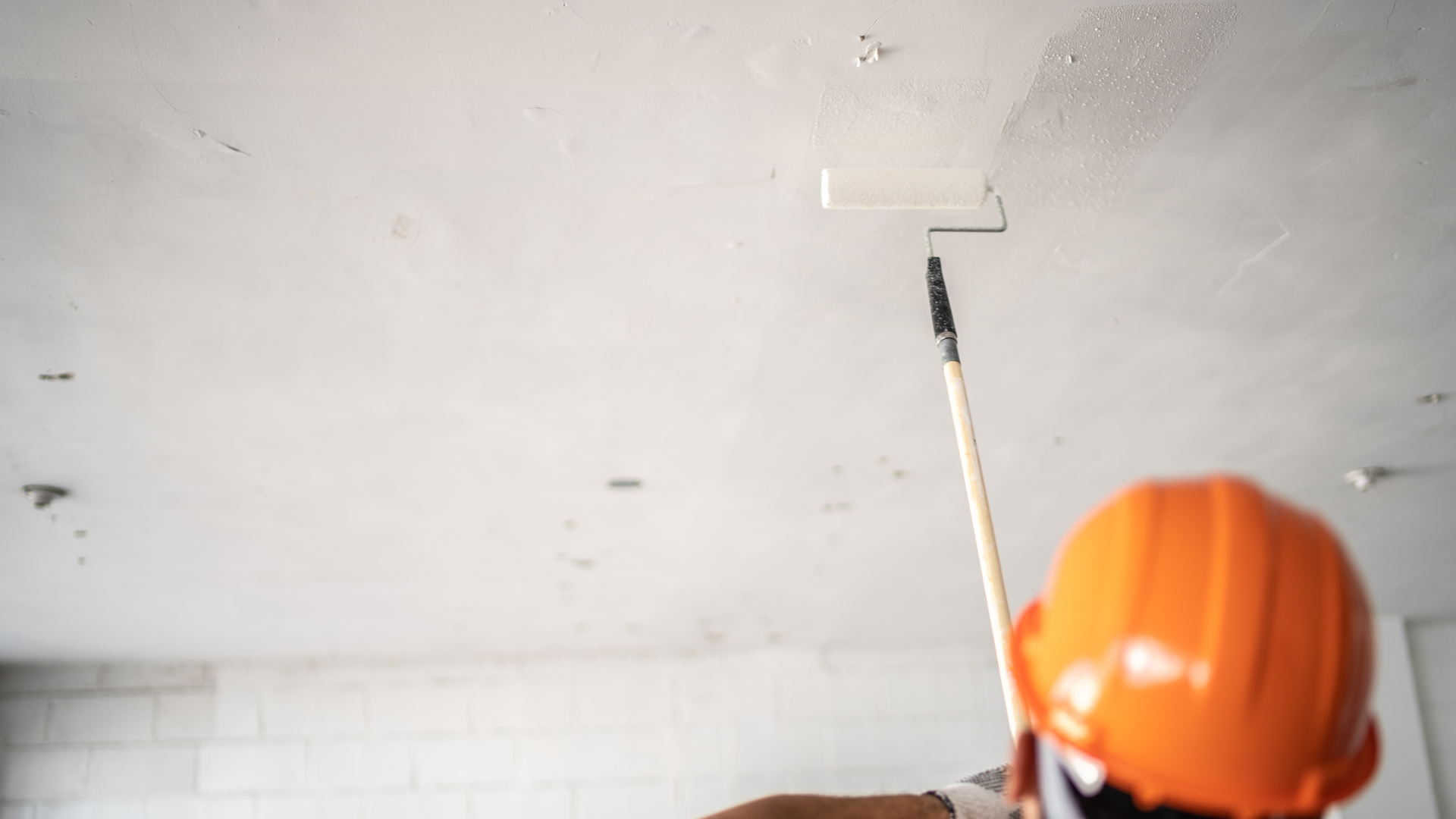 Person in an orange hard hat painting a ceiling with a roller, addressing water stains and preparing for a fresh coat, emphasizing proper ceiling restoration techniques.