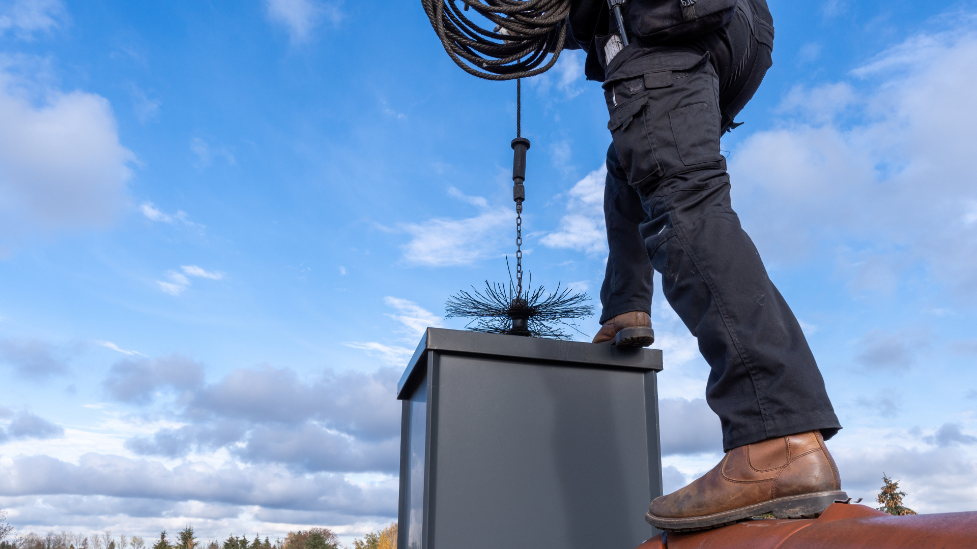 Chimney cleaning process with technician using a brush and rope on chimney cap against a blue sky.