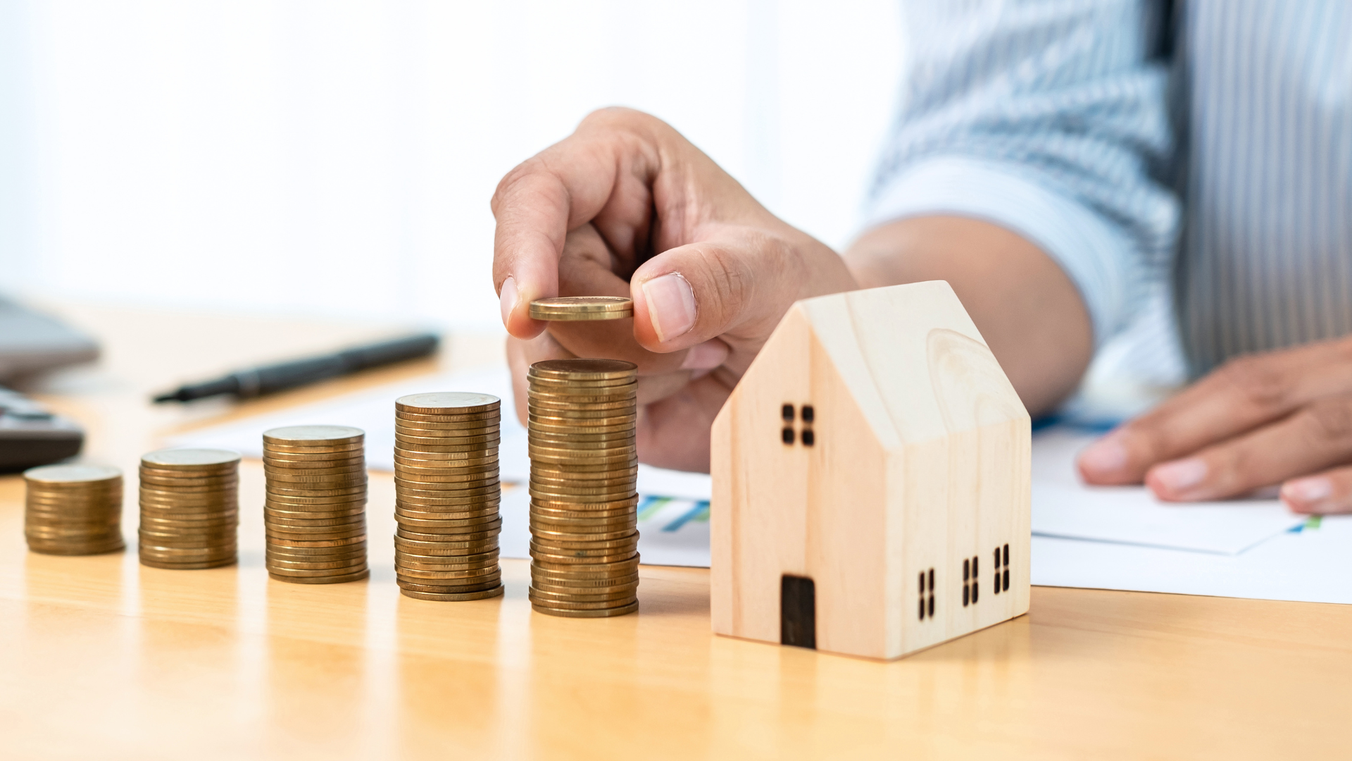Hand placing a coin on a stack of coins next to a wooden house model, symbolizing financial investment in home value enhancement.