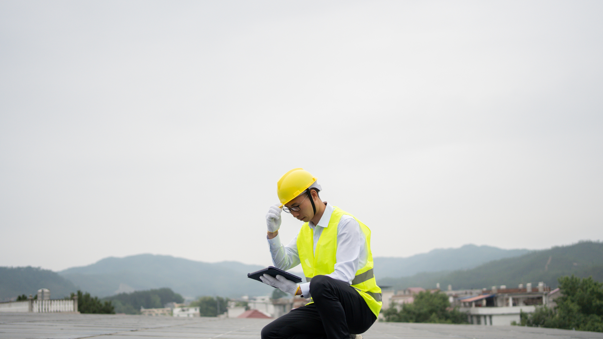 Man in safety gear and yellow hard hat inspecting a roof with a tablet, emphasizing the importance of roof inspections for real estate evaluations.