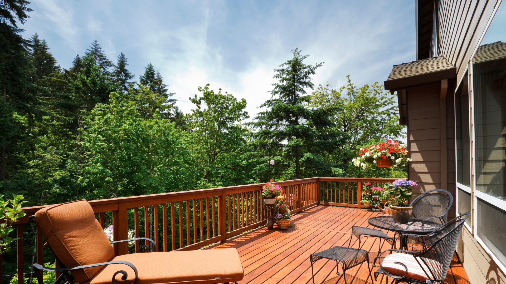 Deck with orange lounge chair and black chairs, surrounded by lush greenery and flower pots, showcasing an inviting outdoor living space ideal for relaxation and gatherings.
