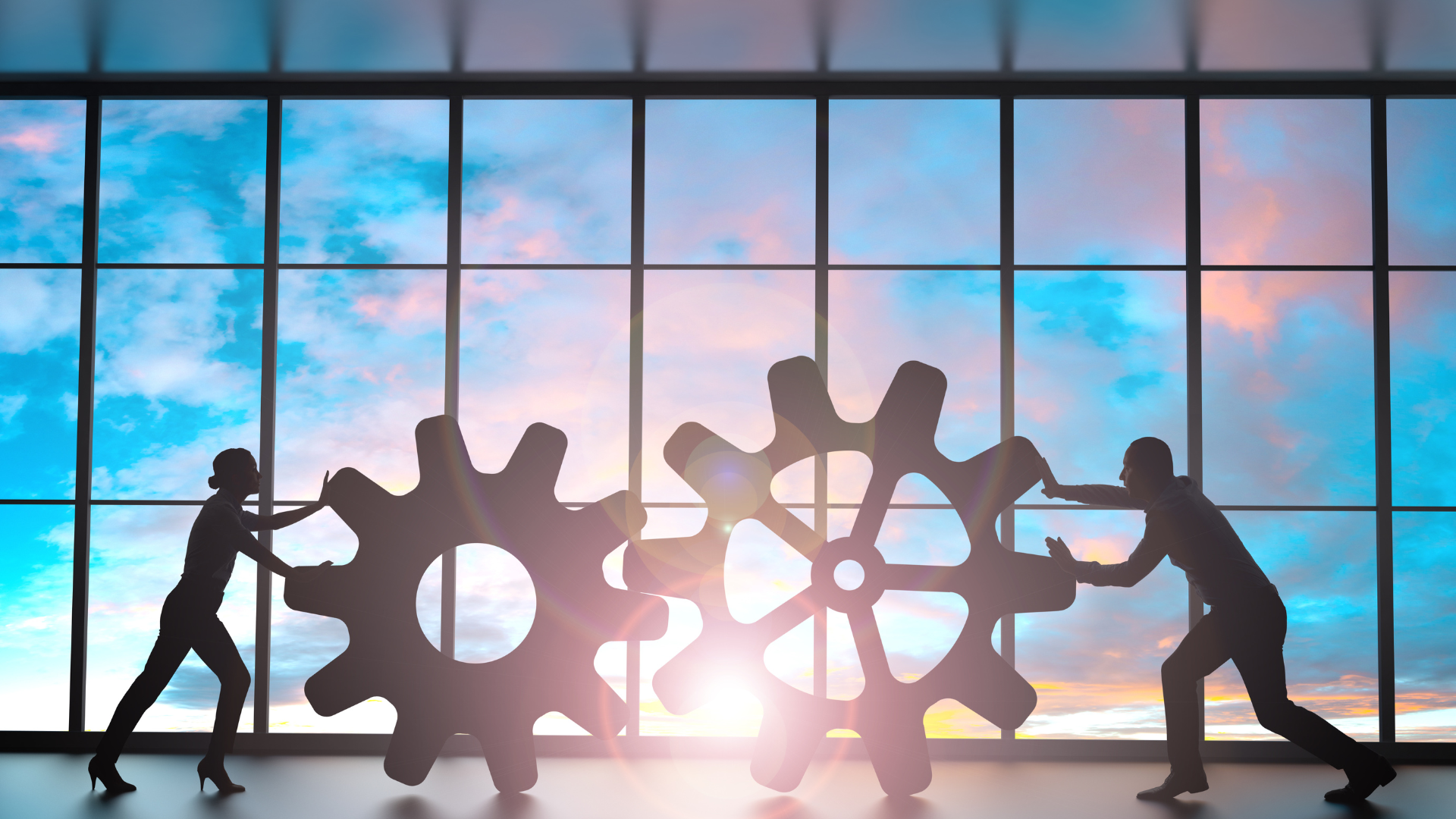 Silhouettes of a man and woman pushing large gears against a backdrop of a colorful sunset through a window, symbolizing teamwork and collaboration in asphalt paving projects.